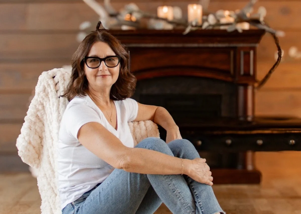 Joyce Jeziorski sitting relaxed in a white tee and jeans in front of a wooden fireplace
